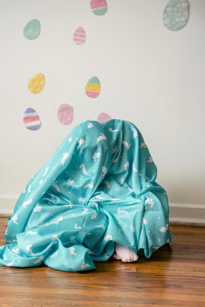 Children playfully hide under a rabbit-decorated blanket with Easter eggs in the background.